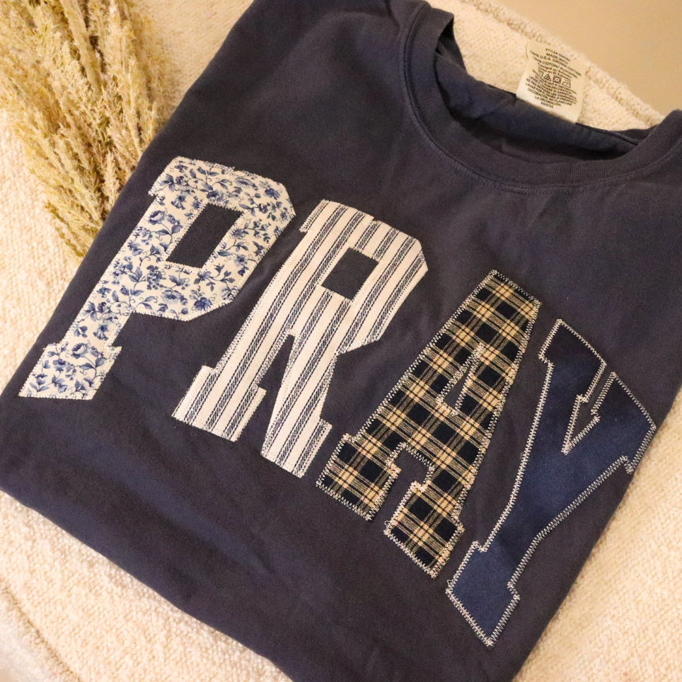 Navy blue t-shirt with 'PRAY' text in various patterns like patchwork.