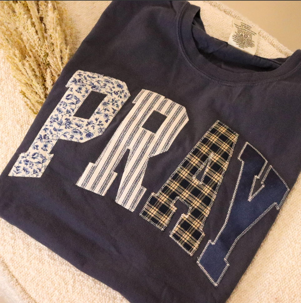 Navy blue t-shirt with 'PRAY' text in various patterns like patchwork.
