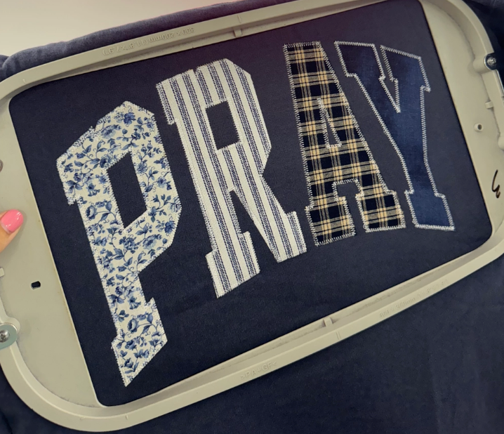 Navy blue t-shirt with 'PRAY' text in various patterns like patchwork.